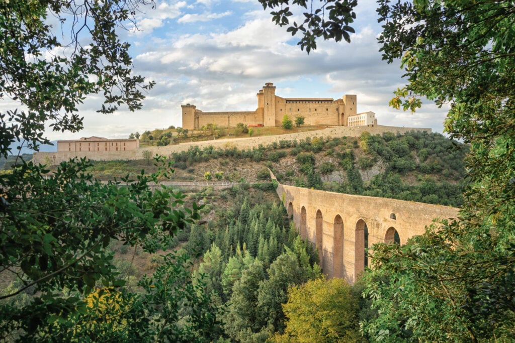 Un grand château se dresse au sommet d'une colline, entouré d'arbres et de verdure. Un ancien aqueduc en pierre aux nombreuses arcades traverse la vallée boisée en contrebas et mène au château. Le ciel est partiellement nuageux. - Karavaniers
