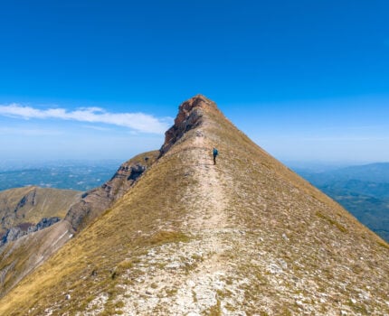 Une personne fait une randonnée le long d'une crête étroite et rocheuse, sous un ciel bleu limpide, avec au loin des collines et des vallées visibles de part et d'autre de la crête. - Karavaniers