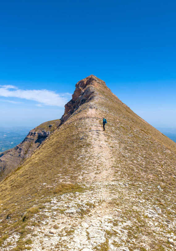 Une personne fait une randonnée le long d'une crête étroite et rocheuse, sous un ciel bleu limpide, avec au loin des collines et des vallées visibles de part et d'autre de la crête. - Karavaniers