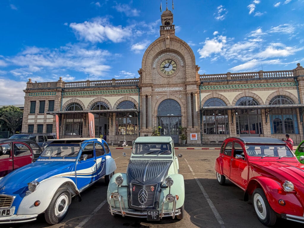 Trois Citroën 2CV d'époque — bleue, verte et rouge — sont garées devant une gare historique en pierre, dotée d'une horloge et de fenêtres cintrées, sous un ciel bleu parsemé de nuages. - Karavaniers