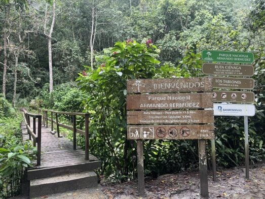 Une passerelle en bois mène à une forêt dense, à côté d'un panneau de randonnée indiquant le parc national Armando Bermúdez, sur lequel figurent les distances des itinéraires et le règlement du parc. - Karavaniers