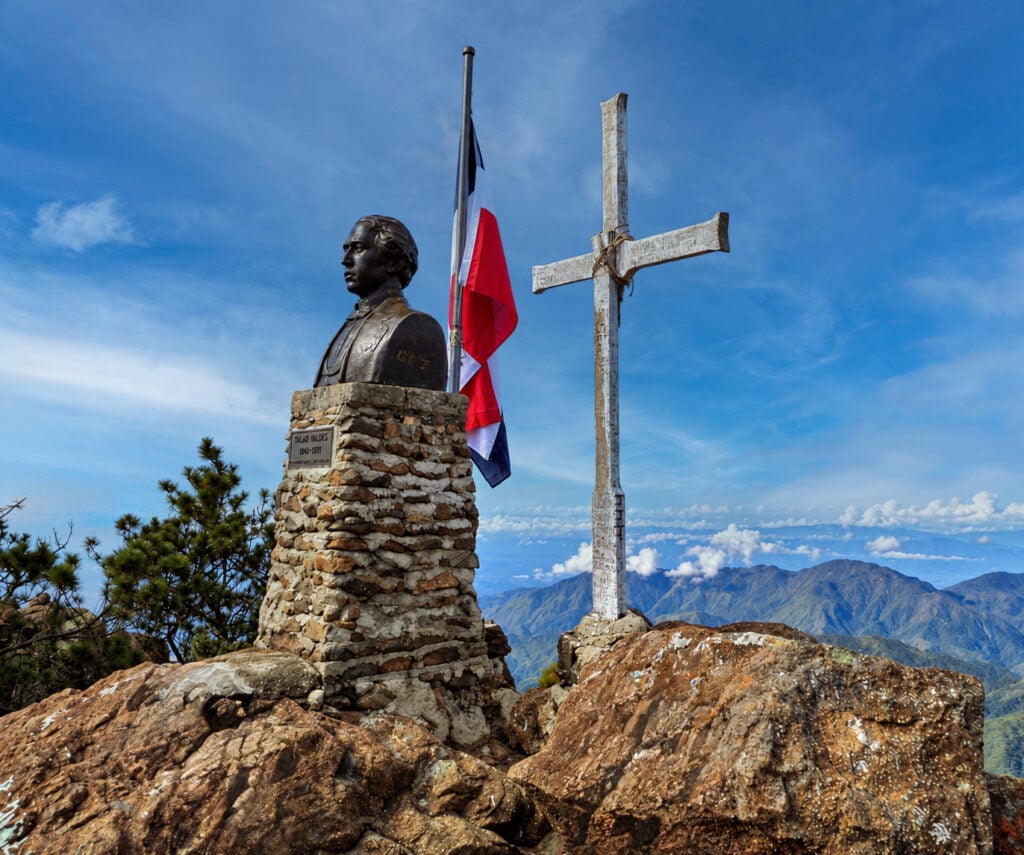 Un buste en bronze de Tirad Valdes posé sur un socle en pierre, à côté d'un drapeau de la République dominicaine en berne et d'une croix en bois patinée par les intempéries, au sommet d'une montagne, lors d'une randonnée en République Dominicaine. - Karavaniers