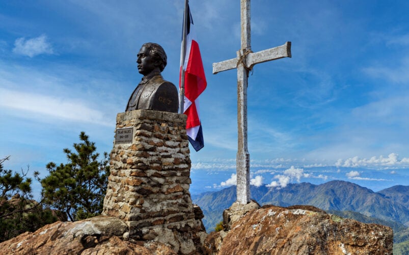 Un buste en bronze de Tirad Valdes posé sur un socle en pierre, à côté d'un drapeau de la République dominicaine en berne et d'une croix en bois patinée par les intempéries, au sommet d'une montagne, lors d'une randonnée en République Dominicaine. - Karavaniers