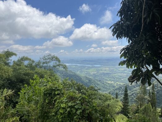 Une vue panoramique depuis une montagne lors d'une randonnée en République Dominicaine, donnant sur une vallée verdoyante et un lac au loin, sous un ciel bleu et nuageux. - Karavaniers