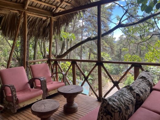 Une terrasse en bois sous un toit de chaume, avec deux chaises roses, deux petites tables en bois et un banc orné de coussins à motifs, donnant sur un sentier de randonnée dans la forêt en République Dominicaine. - Karavaniers