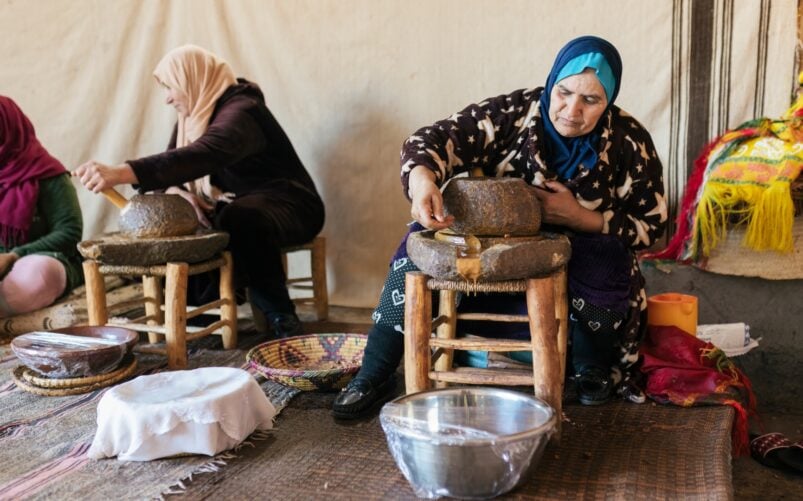 Trois femmes en voyage au Groenland sont assises sur des tabourets bas et moulent du grain à l'aide de moulins à pierre traditionnels à l'intérieur d'une tente. - Karavaniers
