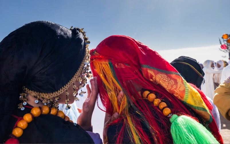 Deux femmes portant des coiffes traditionnelles colorées et des colliers de perles se tiennent côte à côte en plein air lors d'un voyage au Groenland. - Karavaniers